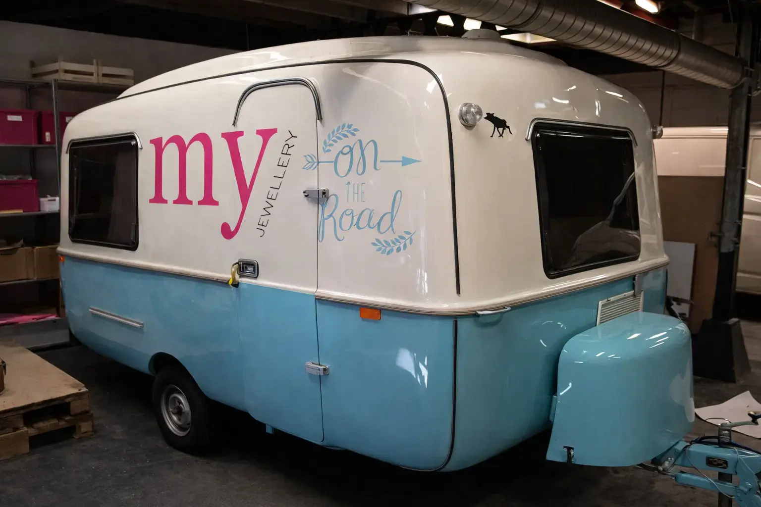 Wit-blauwe caravan met voertuigbelettering in roze en lichtblauw, voorzien van de tekst “my jewellery on the road”, gefotografeerd in een werkplaats.