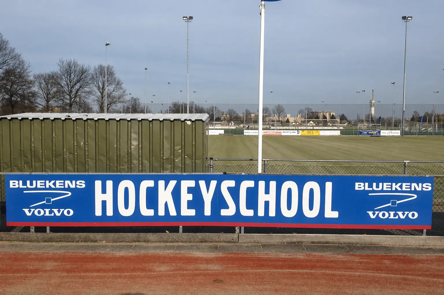 Blauw reclamebord met Bluekens Volvo en HOCKEYSCHOOL langs een omheind hockeyveld.