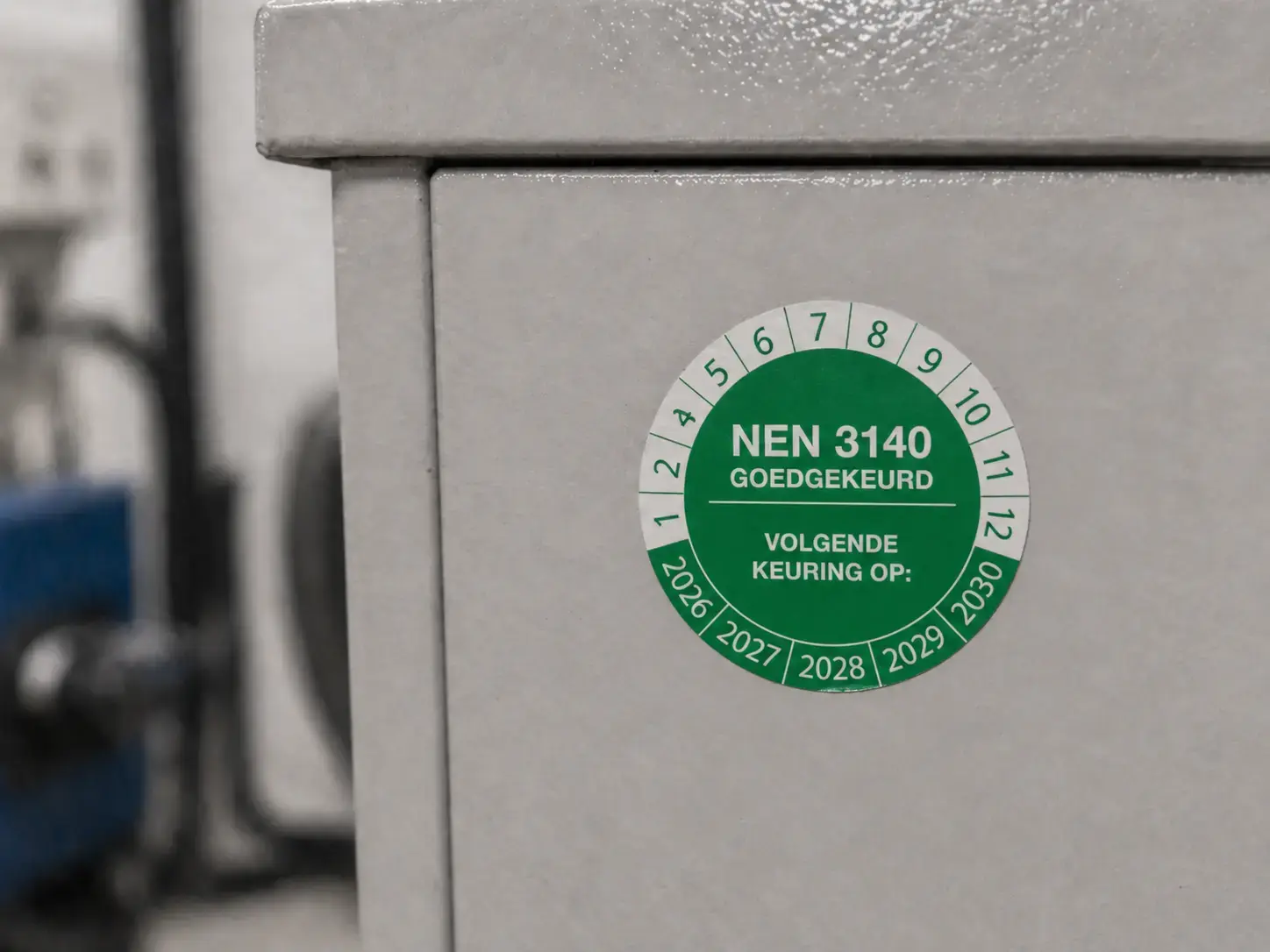 Groene NEN 3140 keuringssticker op een grijze elektrische kast, gebruikt als veiligheidslabel voor goedgekeurde inspectie en volgende keuring.