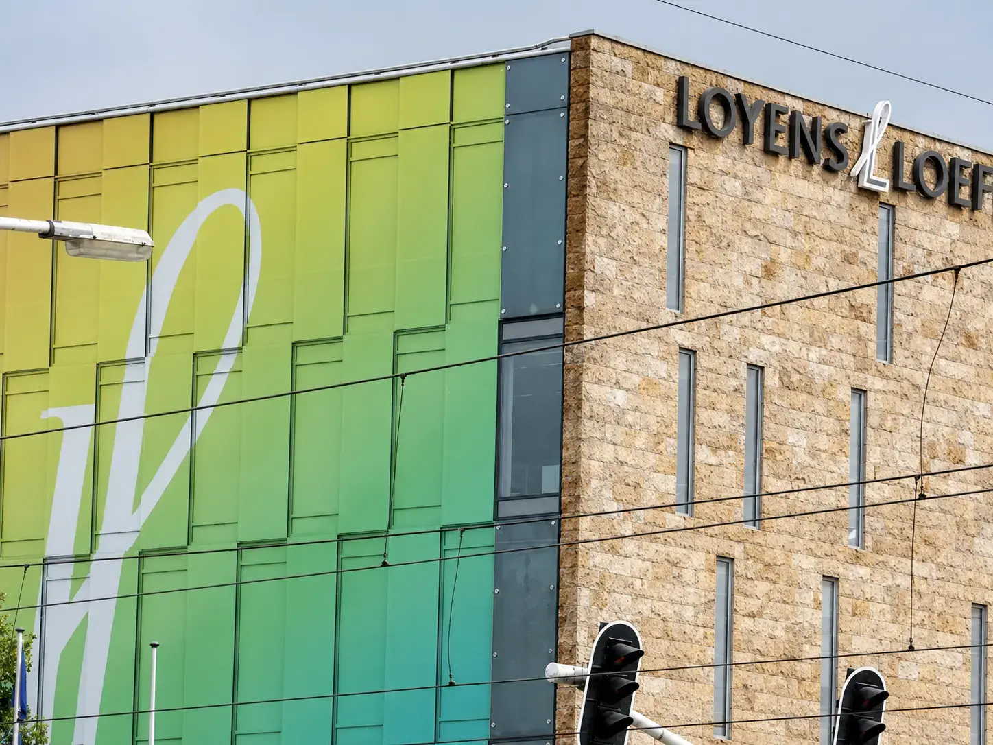 Hoekgevel van een modern kantoorgebouw met zwarte gevelletters "Loyens & Loeff" op de natuurstenen gevel en groen-gele raamfolie met grote witte vorm op de glazen zijgevel.