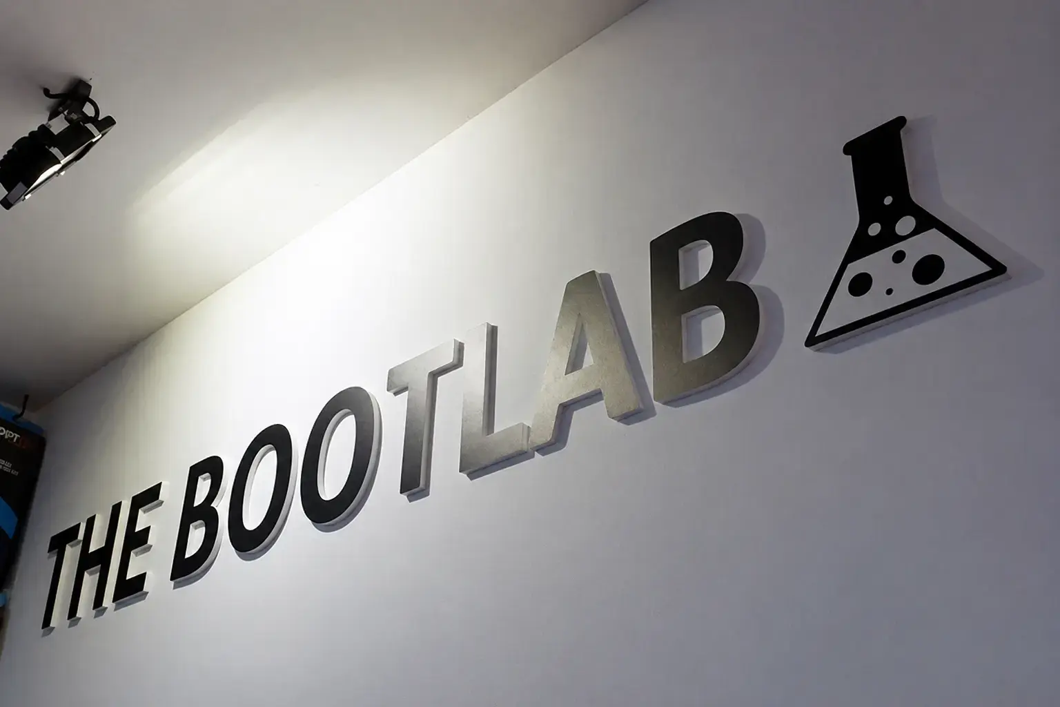 Wandbord met 3D freesletters The Bootlab en een zwart pictogram, gemonteerd op een witte binnenmuur met spotverlichting.