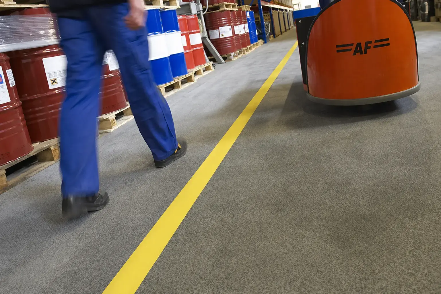 Gele vloerbelijning in een magazijnpad met opslagvaten, medewerker en oranje intern transportvoertuig, toegepast als duidelijke veiligheidsmarkering en looproute.