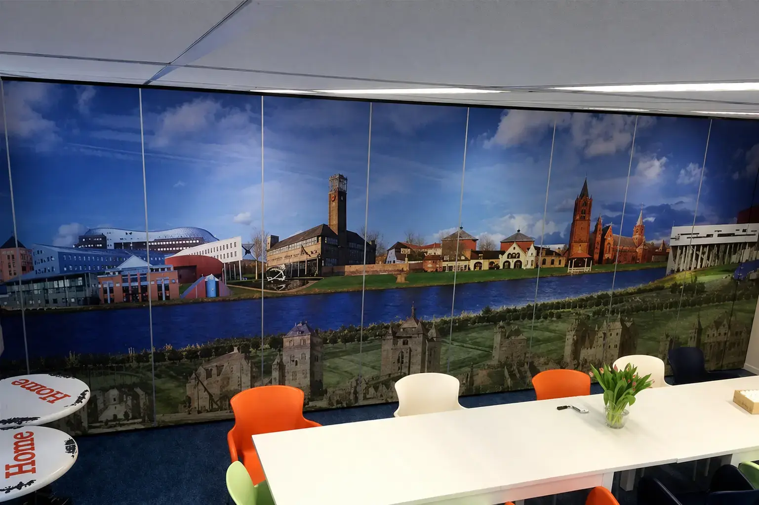 Vergaderruimte met kamerhoge wandprint van een stadsgezicht, toegepast als decoratieve muurvisual en interieurbelettering achter een lange tafel.
