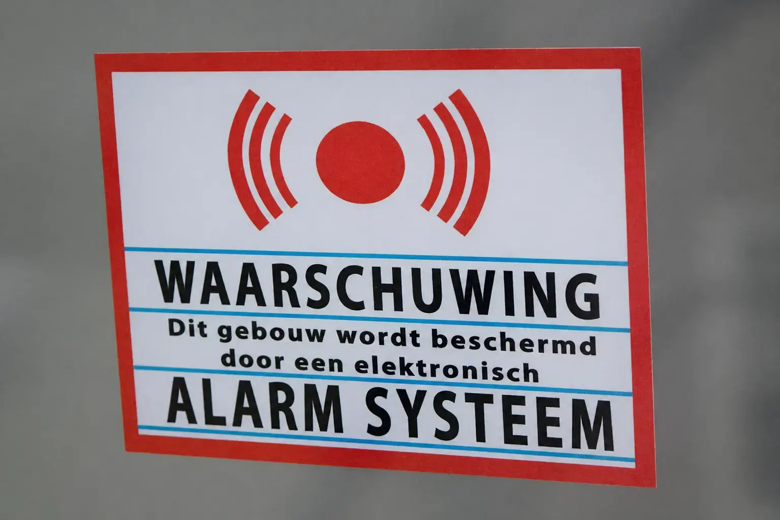 Waarschuwingssticker met rood alarmsymbool en tekst over elektronisch alarmsysteem, aangebracht op een grijze ondergrond als beveiligingssignalering.