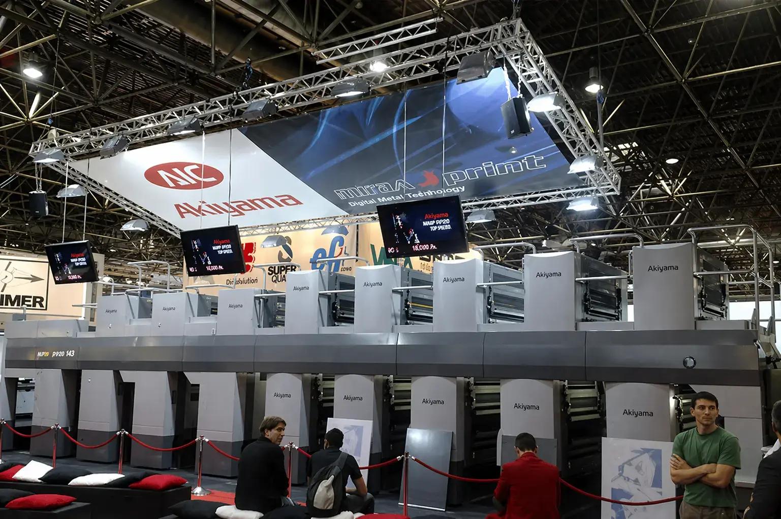Groot hangend beursbanner in trussconstructie met verlichting boven een Akiyama drukpers op een vakbeurs.