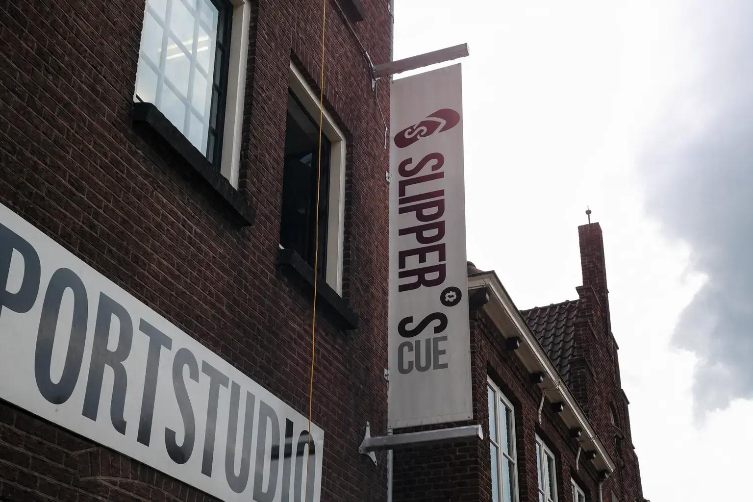 Verticale gevelbanner met meerkleurige belettering aan een bakstenen pand, naast een gedeeltelijk zichtbaar wit gevelbord met grote zwarte letters.