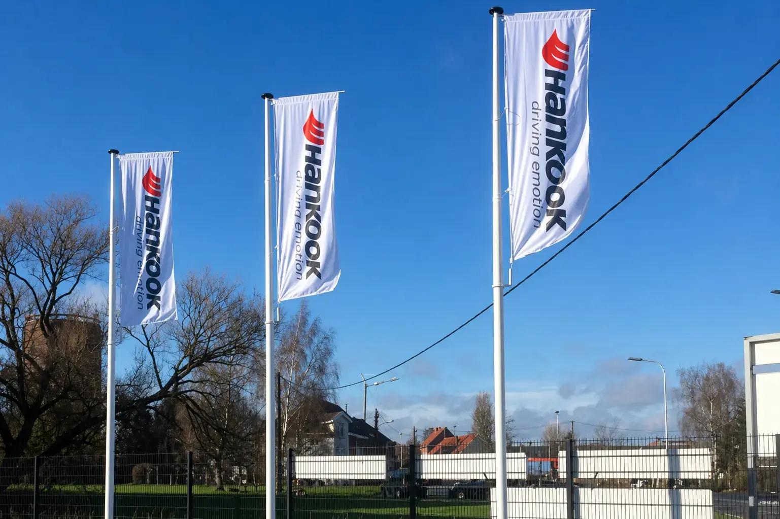 Drie witte mastvlaggen met Hankook-logo en verticale belettering, geplaatst op hoge vlaggenmasten op een bedrijventerrein onder een heldere blauwe lucht.