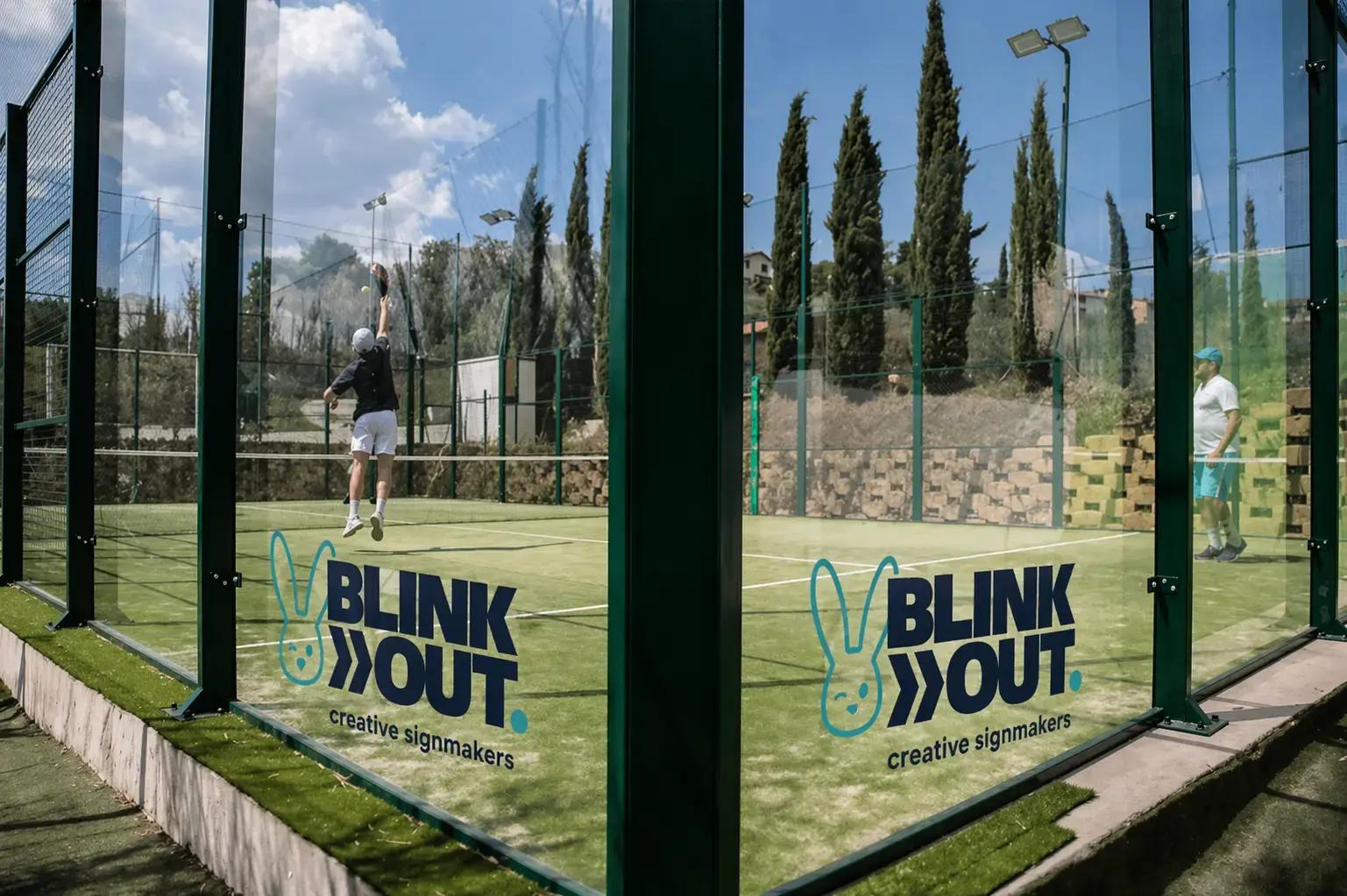 Glaswand van een padelbaan met Blink Out creative signmakers-logo in turquoise en donkerblauw, aangebracht als transparante raamsticker op sportbeglazing.