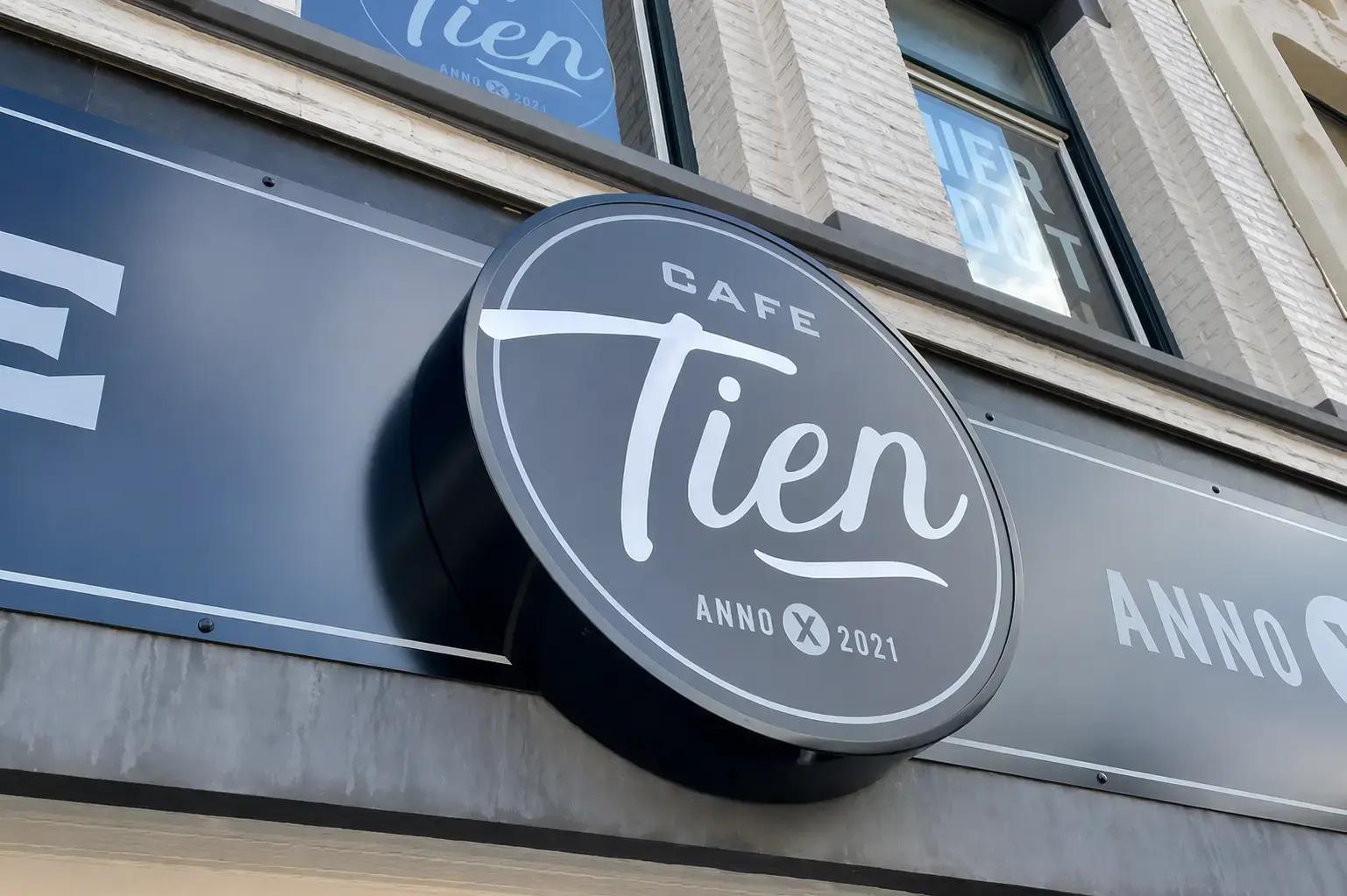Rond donkerblauw gevelbord van Café Tien met witte belettering en logo, gemonteerd als uithangbord aan de voorgevel van een horecapand.