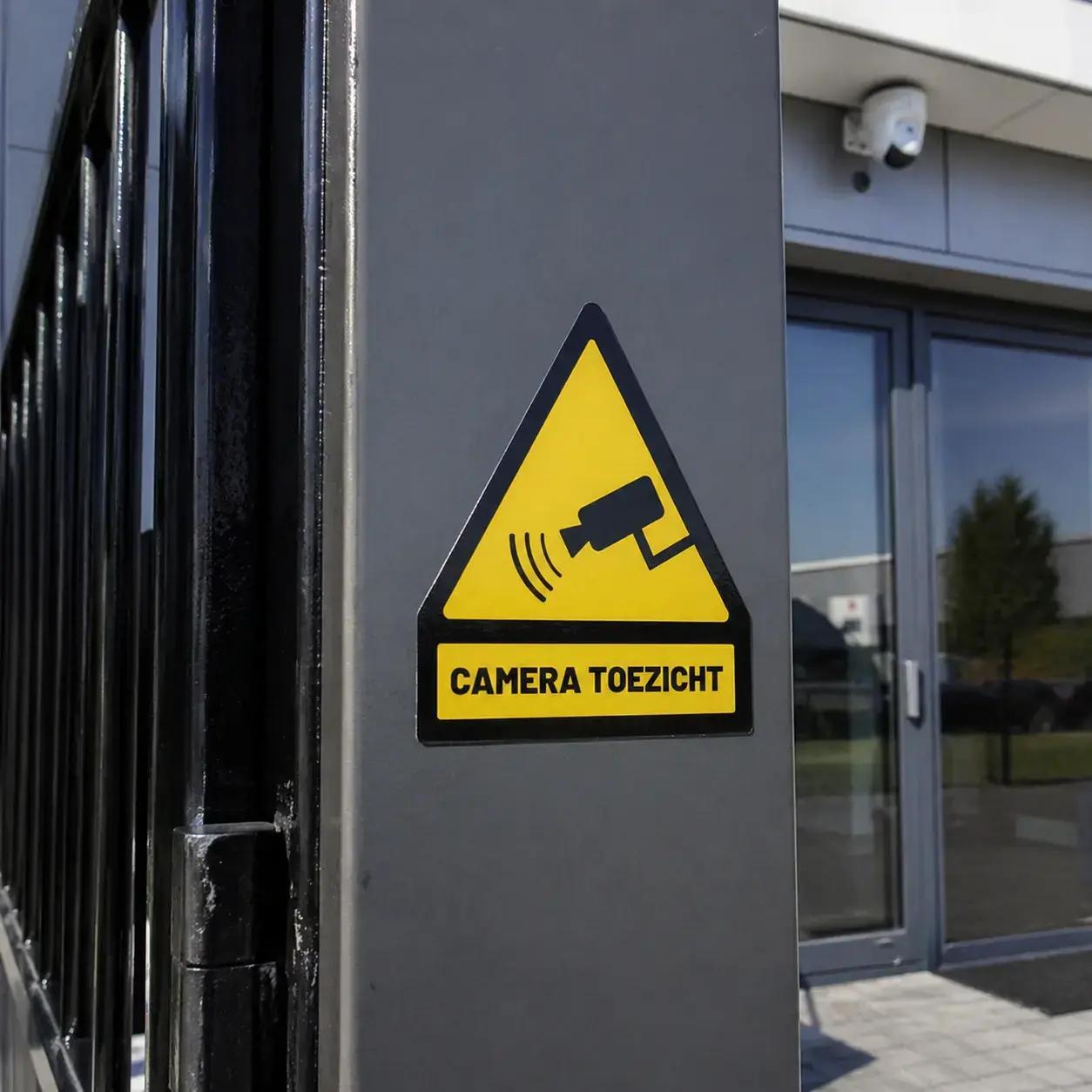 Gele waarschuwingssticker voor cameratoezicht op een grijze buitenzuil bij een toegangspoort en bedrijfsingang.