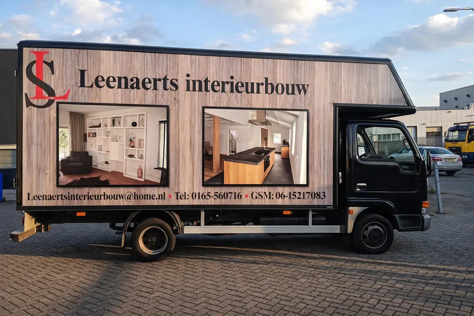 Zwarte bedrijfswagen met full-color voertuigbelettering voor Leenaerts interieurbouw, voorzien van een houtlook wrap, groot logo, zwarte geveltekst en twee interieurfoto’s op de zijwand.