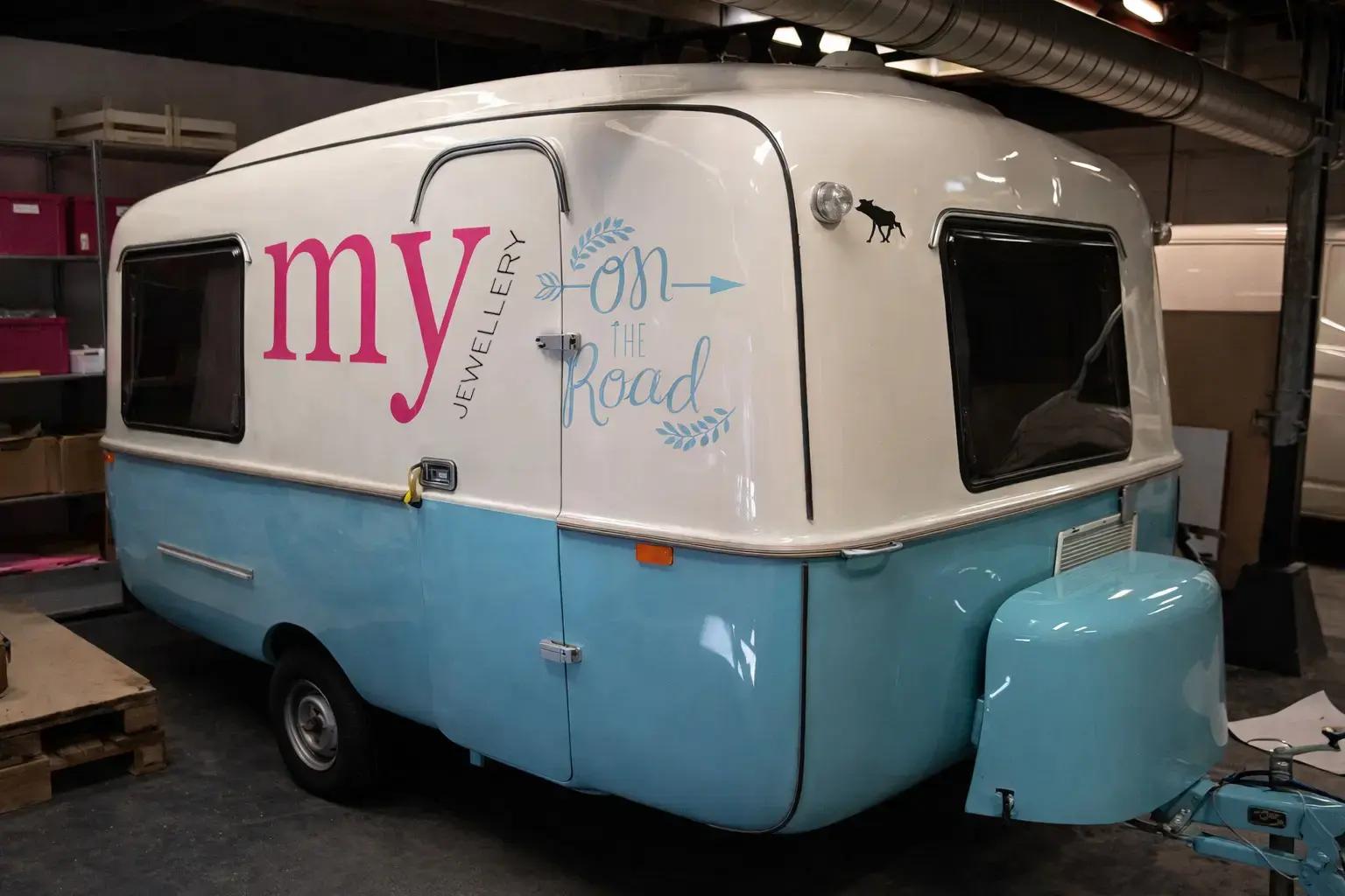 Wit-blauwe caravan met voertuigbelettering in roze en lichtblauw, voorzien van de tekst “my jewellery on the road”, gefotografeerd in een werkplaats.