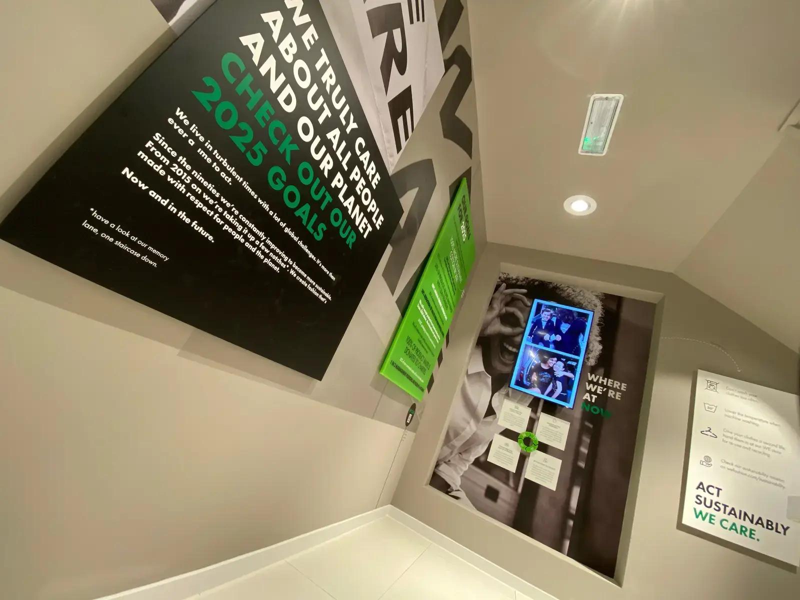 Hoek van een winkelinterieur met grote wandpanelen, interieurbelettering en digitale display over duurzaamheid, waaronder de teksten "CHECK OUT OUR 2025 GOALS", "WHERE WE’RE AT NOW" en "ACT SUSTAINABLY WE CARE."