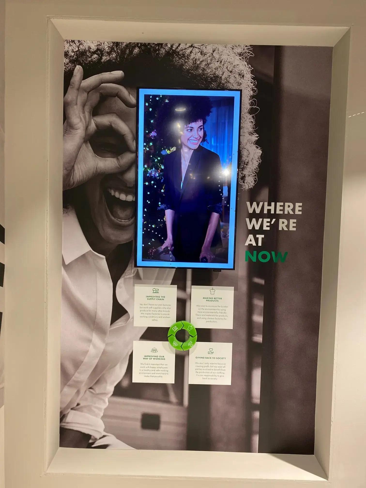 Ingebouwde wandvisual met zwart-wit fotoprint, digitale display en interieurbelettering met de tekst "WHERE WE’RE AT NOW" als informatieve retailpresentatie.