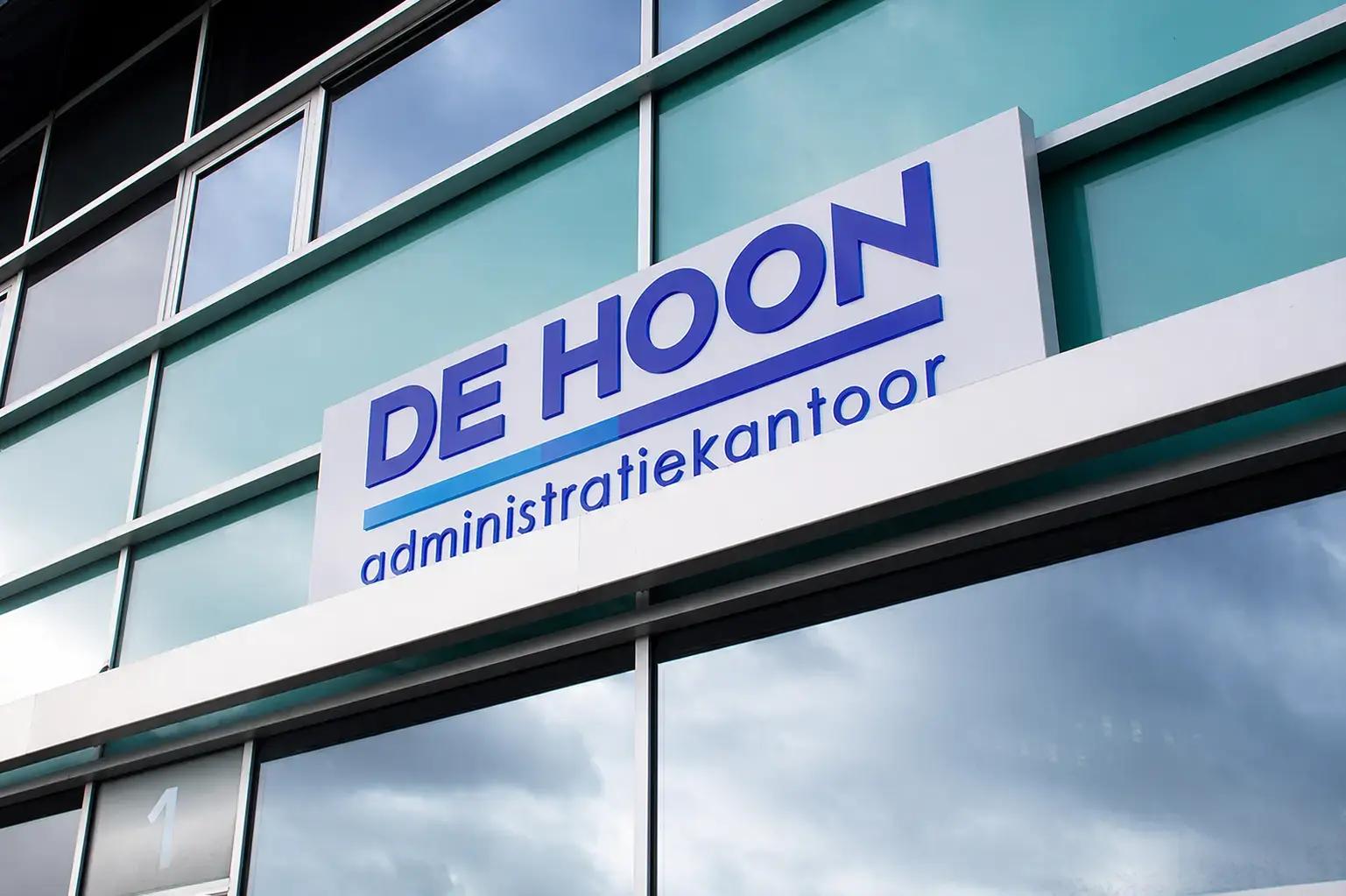 Wit gevelbord met blauwe belettering van DE HOON administratiekantoor, gemonteerd op een moderne glazen gevel boven de entree.