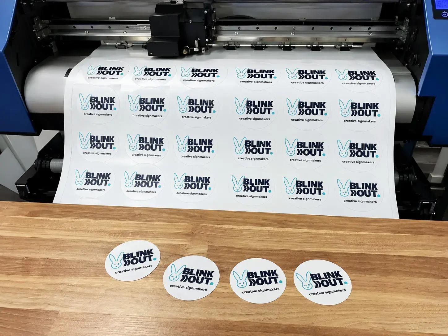 Grootformaat printer die een vel met ronde BLINK OUT creative signmakers-logo’s print, met enkele losse cirkelprints op een houten werkblad ervoor.
