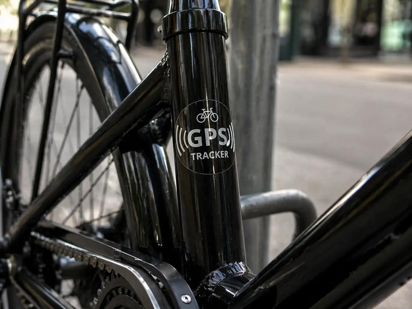 Transparante GPS tracker sticker met wit pictogram en tekst op het zwarte frame van een fiets, toegepast als beveiligingslabel tegen diefstal.