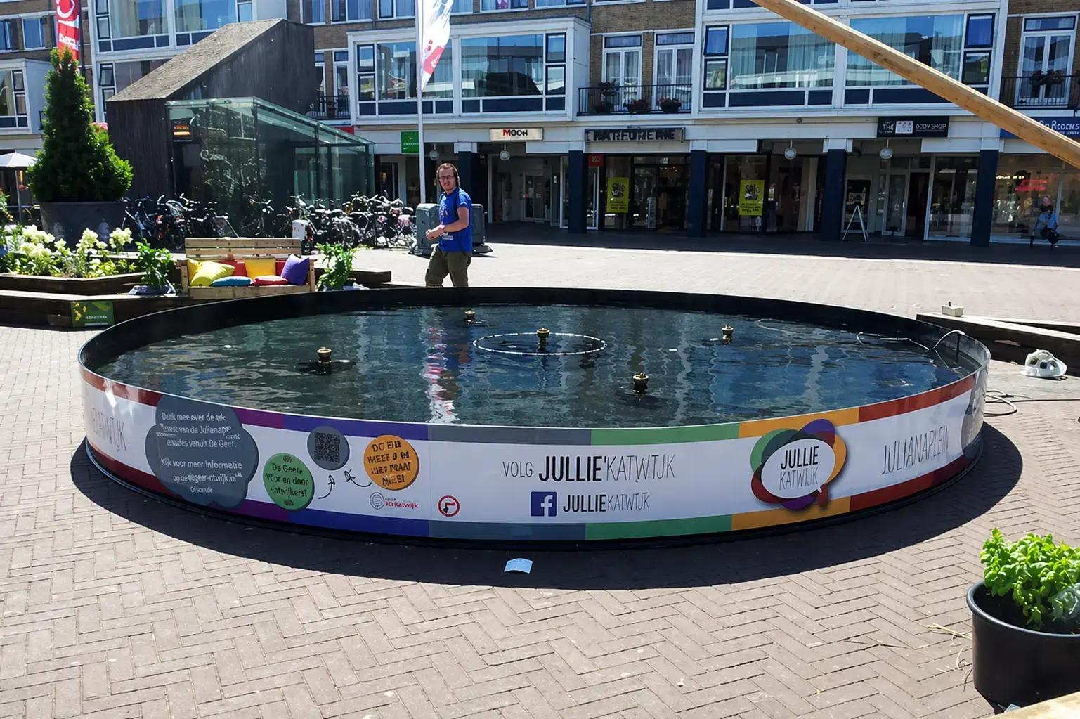 Full colour foliebelettering met Jullie Katwijk op de ronde ombouw van een fontein in een winkelstraat.