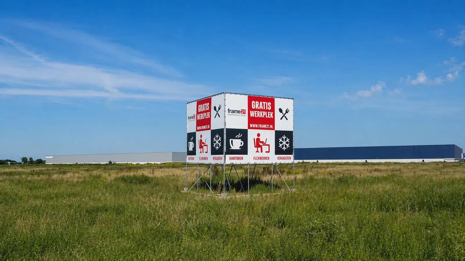 Groot vrijstaand bouwbord met frame21, GRATIS WERKPLEK en pictogrammen voor kantoren, flexwerken en vergaderen op een metalen frame in een grasveld.