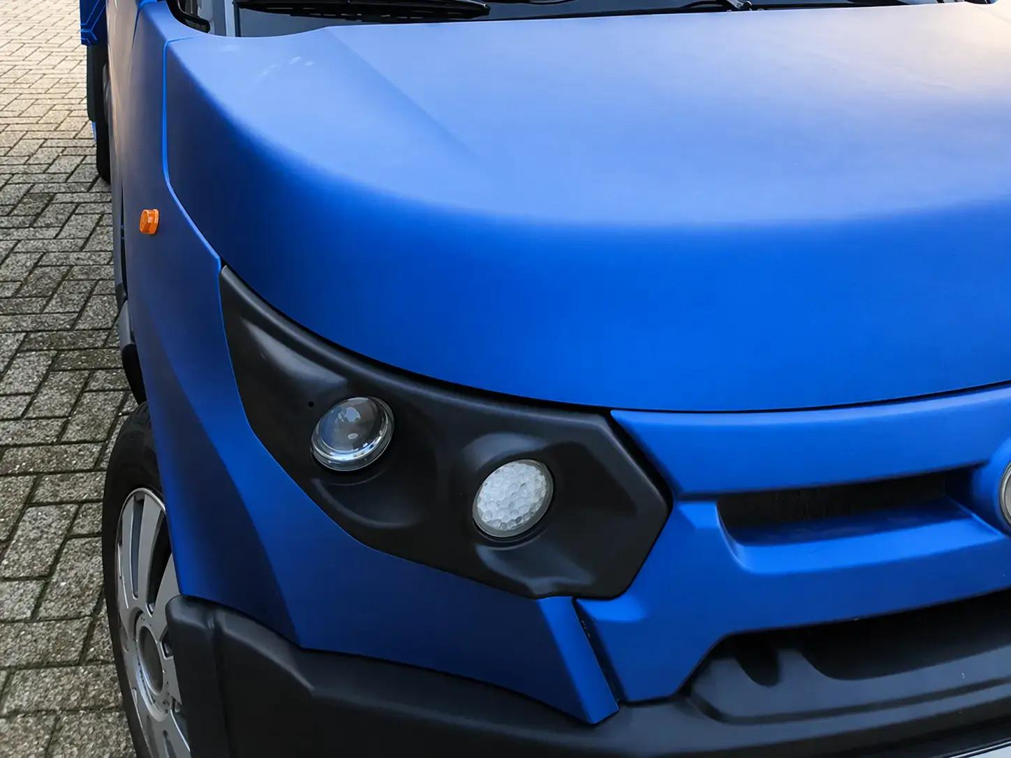 Close-up van de voorzijde van een compact voertuig met matblauwe carwrap, zwarte kunststof accenten en ronde koplampen op straatstenen.