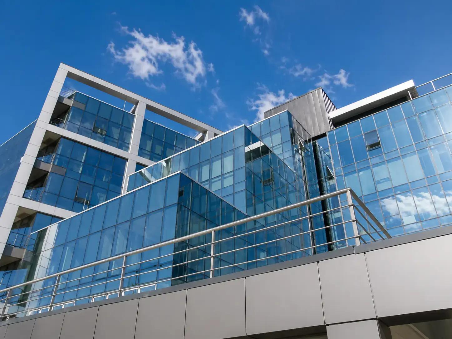 Moderne kantoorgevel met blauwe glaspartijen, reflecterende raamfolie en metalen gevelpanelen onder een heldere lucht.