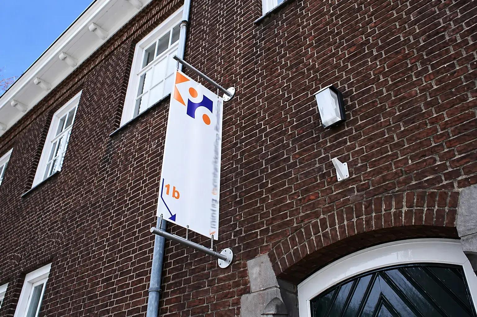 Verticale wayfinding-banner met logo, oranje huisnummer 1b en pijl, gemonteerd aan een bakstenen gevel naast een entree.
