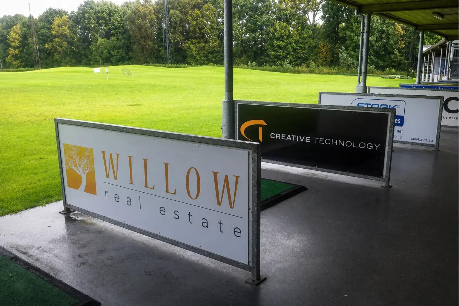 Reclameborden in metalen frames met Willow real estate en Creative Technology langs een overdekte golfafslagplaats bij een groen sportveld.