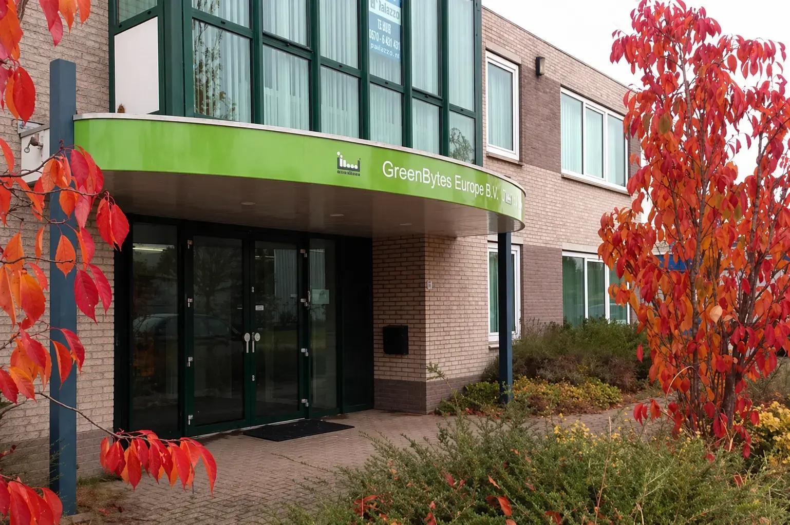 Moderne kantoorentree met groene gevelluifel en witte belettering van GreenBytes Europe B.V. boven glazen deuren, omgeven door herfstkleurige beplanting.
