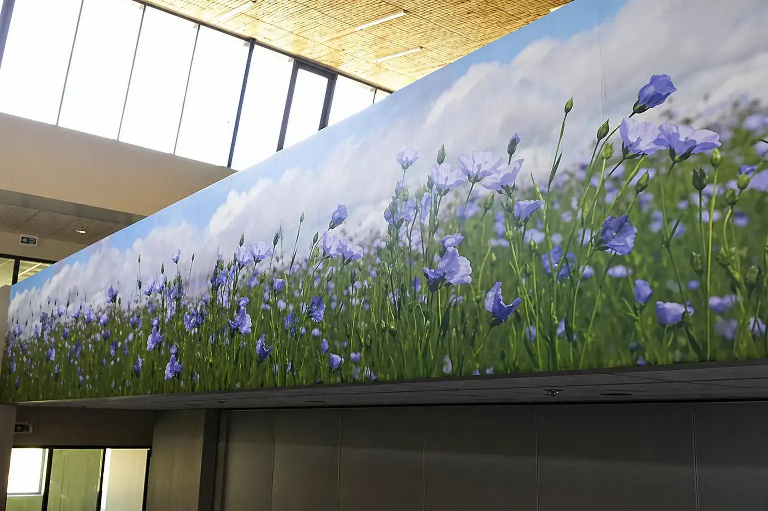Kamerbrede wandprint met blauwe bloemen en wolkenlucht, strak gemonteerd als decoratieve interieurvisual op een hoge binnenwand.