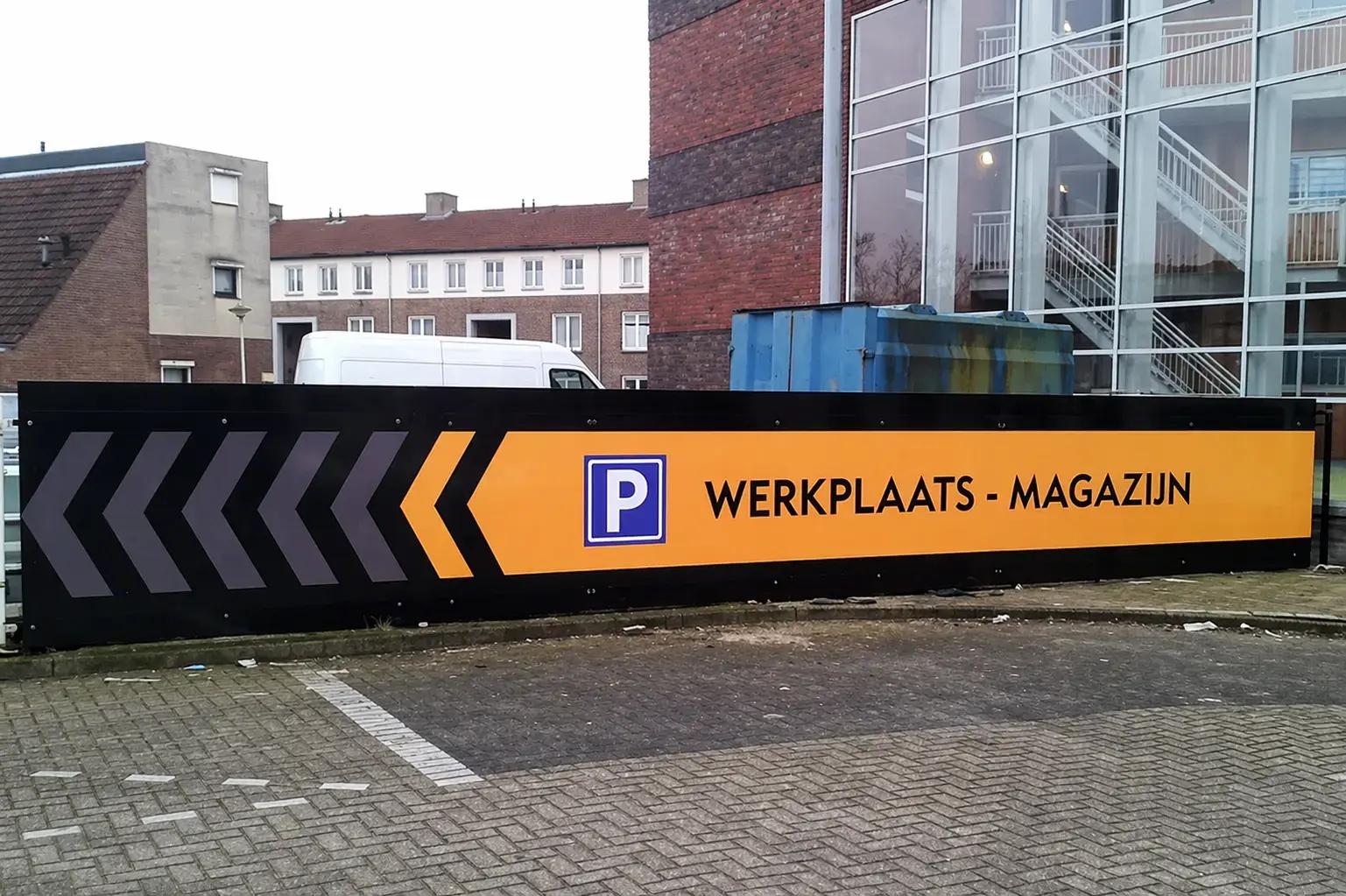 Groot zwart-geel wayfinding-bord met parkeerpictogram, linkerpijlen en de tekst WERKPLAATS - MAGAZIJN, geplaatst langs een parkeerterrein bij een gebouw.