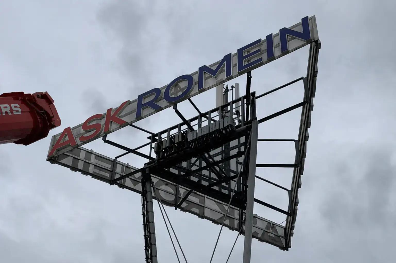 Grote hoog gemonteerde constructie met contourletters ASK ROMEIN in rood en blauw op een metalen frame, gefotografeerd tegen een bewolkte lucht.