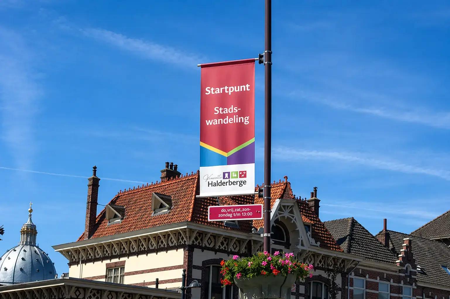 Rode straatbanner aan een lantaarnpaal met Startpunt Stadswandeling Gemeente Halderberge bij historische gebouwen onder een blauwe lucht.