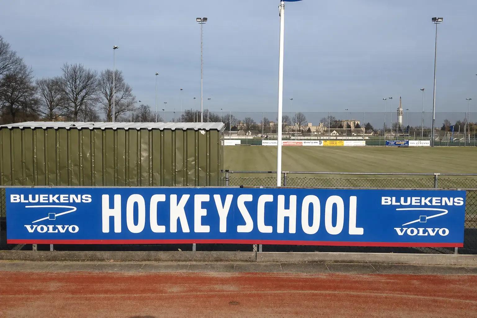 Blauw reclamebord met Bluekens Volvo en HOCKEYSCHOOL langs een omheind hockeyveld.