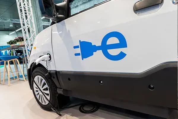 Close-up van een witte elektrische bedrijfswagen aan de laadkabel, met blauwe e-bestickering op het portier en zichtbaar laadpunt tijdens een beurspresentatie.
