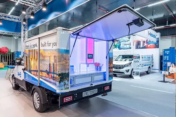 Elektrische StreetScooter bakwagen op een beursstand met opengeklapte laadbak als display, verlichte binnenzijde en full-color voertuigbelettering met stadsbeeld op het zijpaneel.