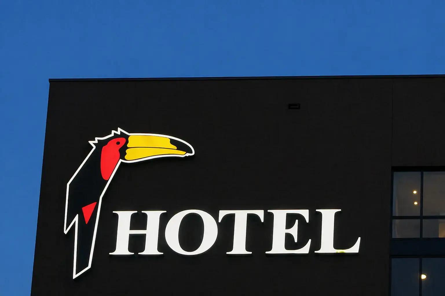 Verlichte gevelreclame met het woord HOTEL en een gestileerde vogelkop op een donkere hotelgevel bij avond.
