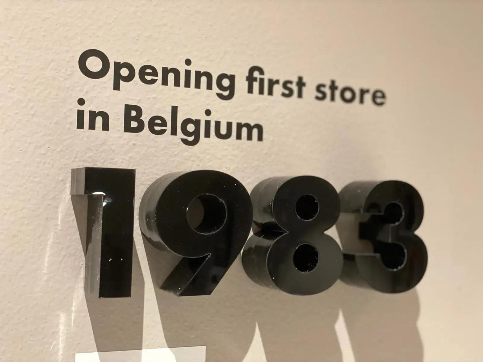 Wandbelettering met zwarte tekst "Opening first store in Belgium" en glanzende 3D cijfers "1983" als typografische interieurcommunicatie op een lichte binnenwand.