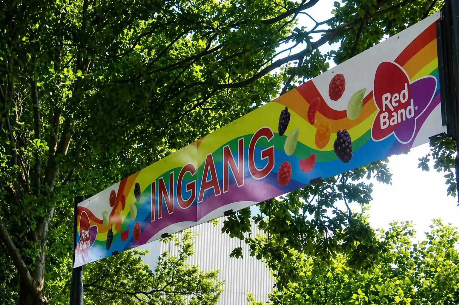 Kleurrijk entreebanner met Red Band logo, snoepafbeeldingen en de tekst INGANG gemonteerd tussen bomen.
