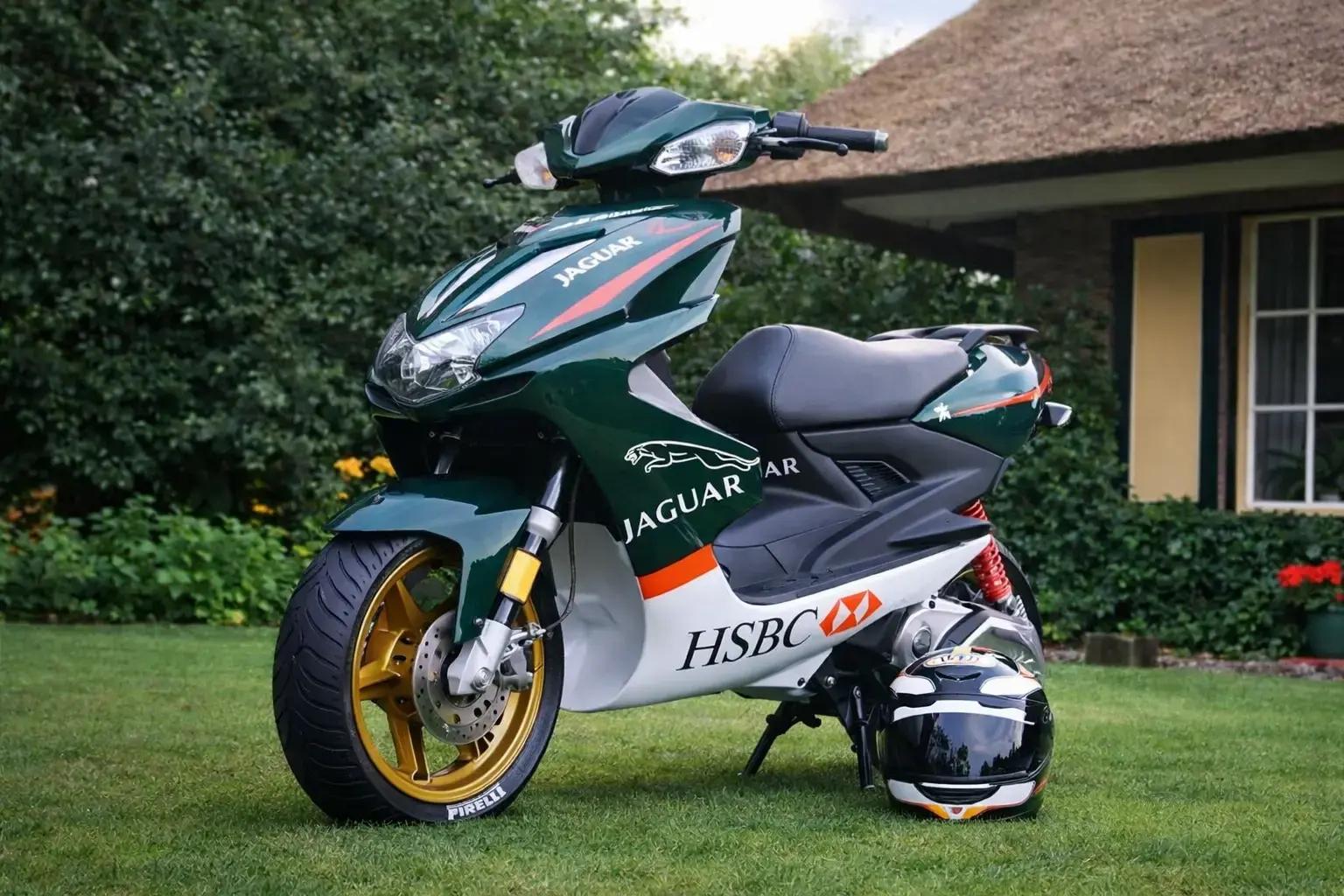 Donkergroene scooter met opvallende voertuigbelettering, Jaguar- en HSBC-logo’s op de kuip, goudkleurige velgen en een helm op het gras voor een woning.