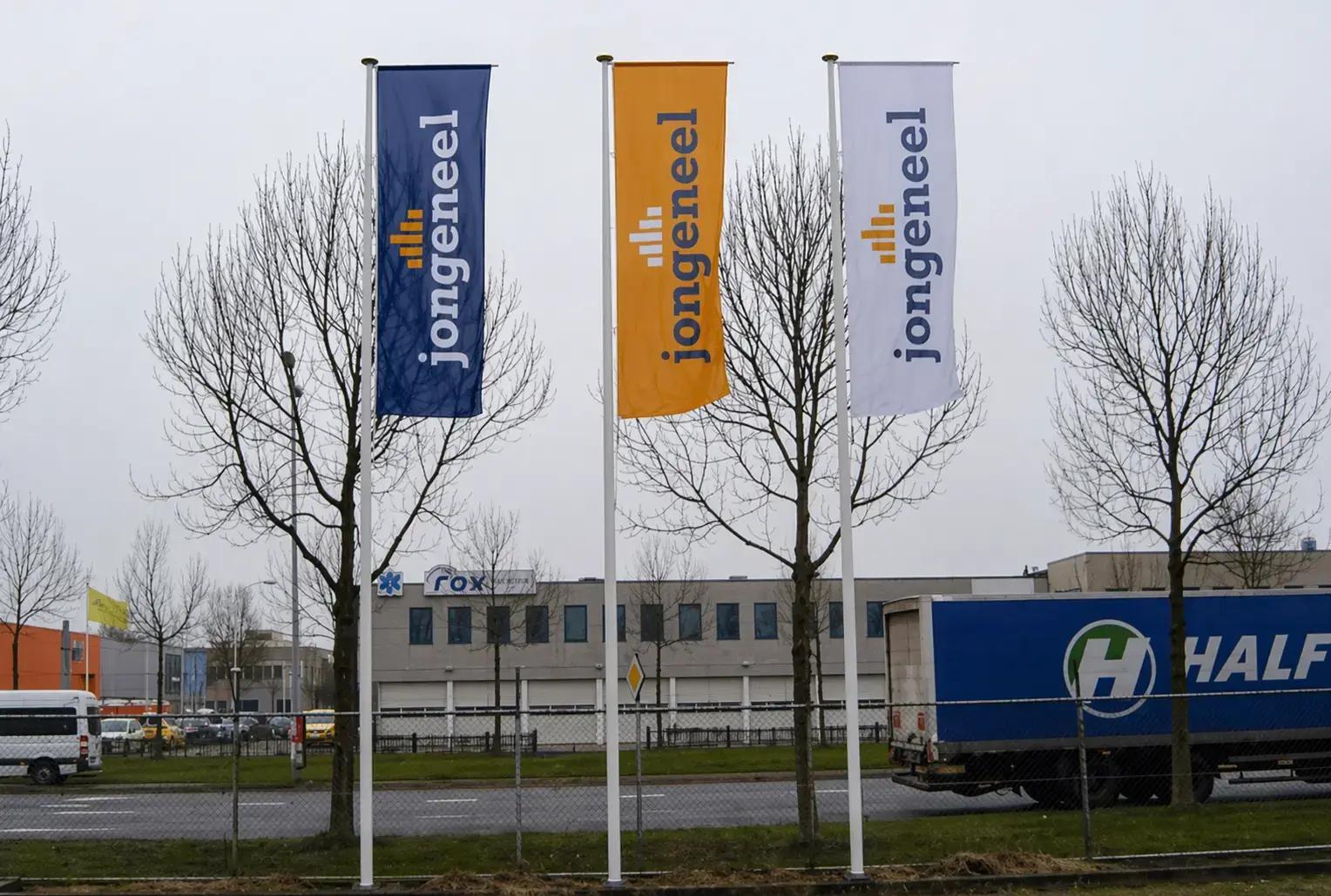 Drie verticale baniervlaggen met Jongeneel-logo in blauw, oranje en wit aan vlaggenmasten langs een bedrijventerrein.