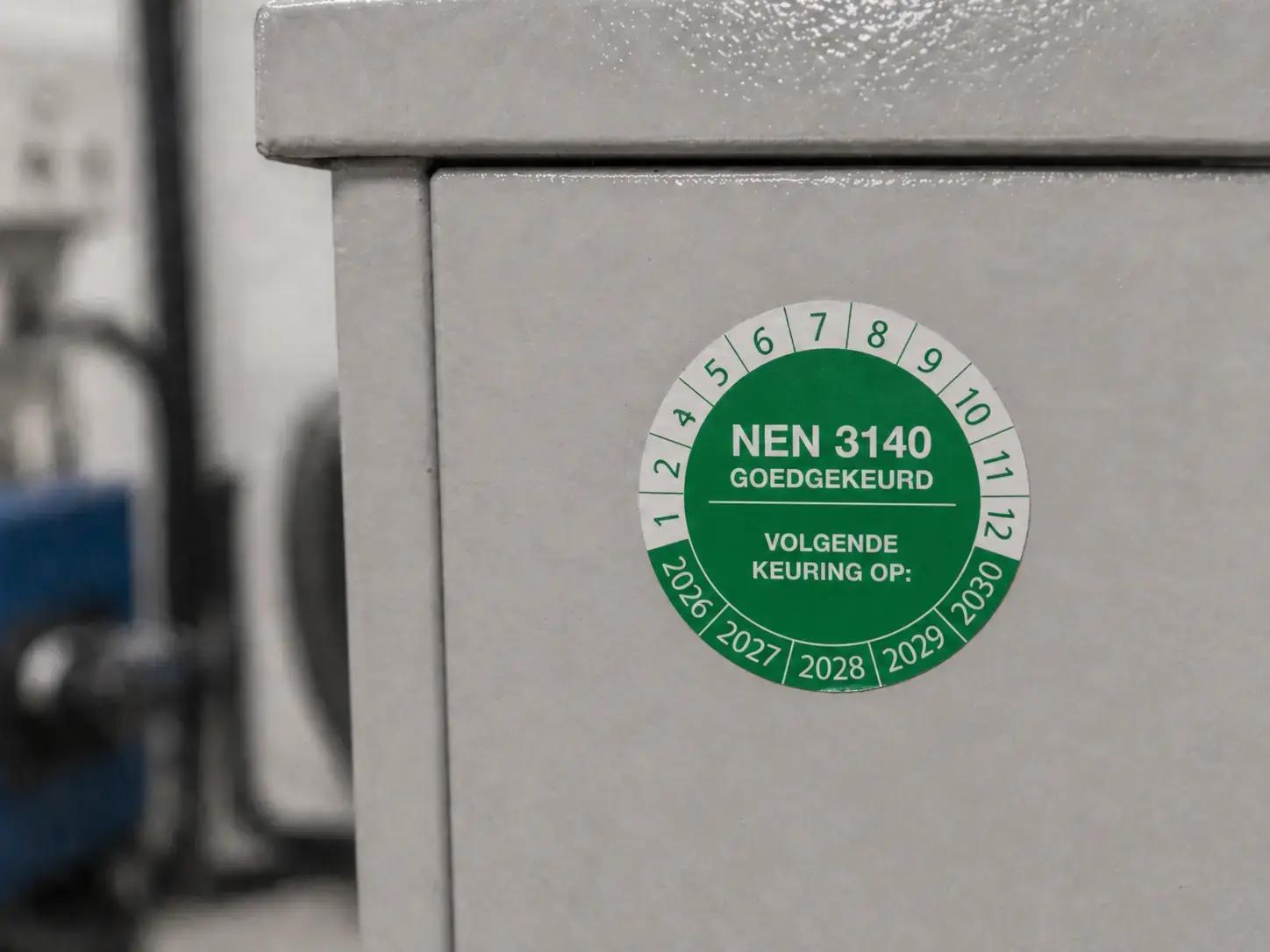 Groene NEN 3140 keuringssticker op een grijze elektrische kast, gebruikt als veiligheidslabel voor goedgekeurde inspectie en volgende keuring.