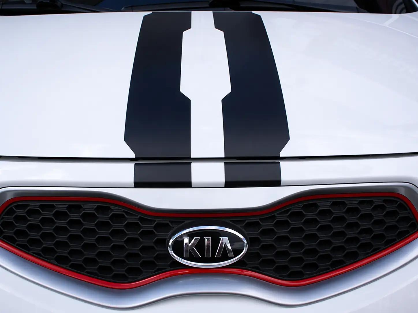 Close-up van de voorzijde van een witte Kia met zwarte striping op de motorkap en rode accenten rond de grille, geschikt als voorbeeld van voertuigbelettering en car wrapping.