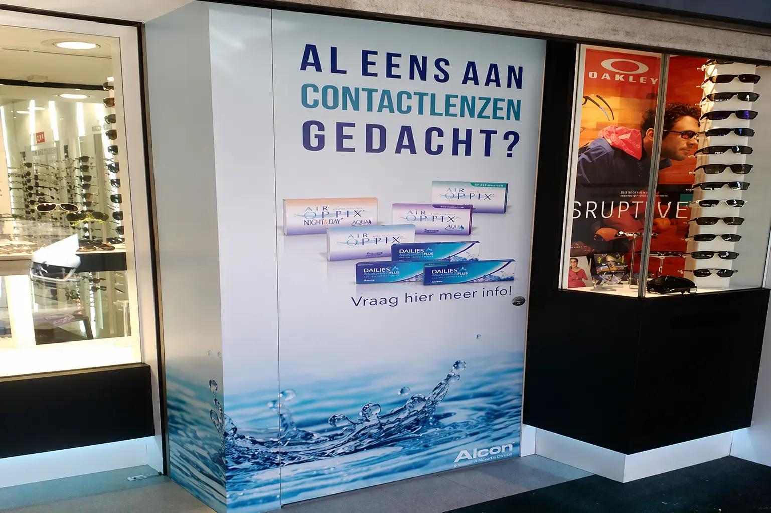 Grote full-colour wandreclame in een optiekzaak met contactlenzenpromotie, waterafbeelding en zichtbare productverpakkingen, naast een etalage met zonnebrillen en Oakley-display.