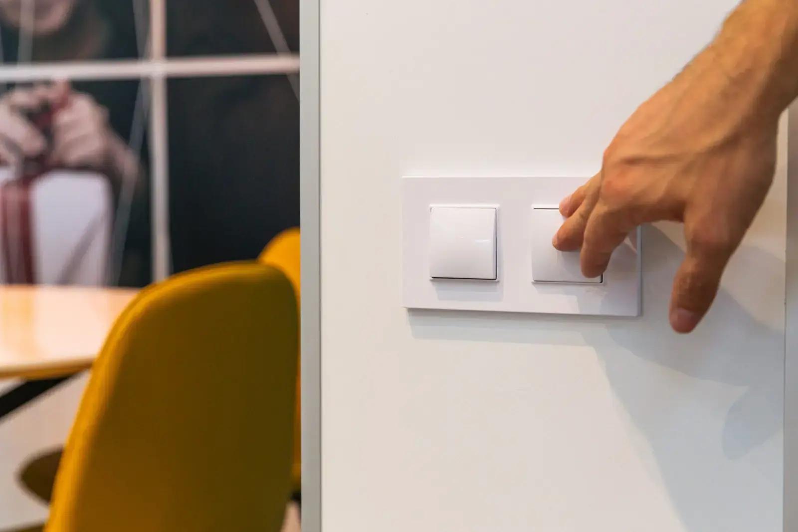 Hand die een witte lichtschakelaar op een wand indrukt in een moderne kantoorruimte met gele stoel op de achtergrond.