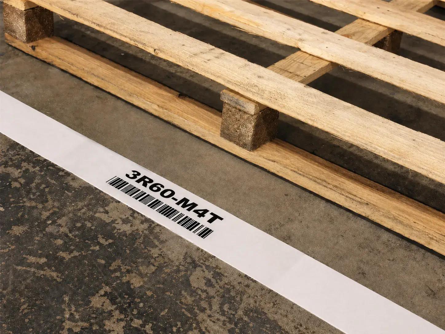 Witte vloermarkering met barcode en code 3R60-M4T naast houten pallets op een magazijnvloer, toegepast als duurzame locatieaanduiding voor logistiek en voorraadbeheer.