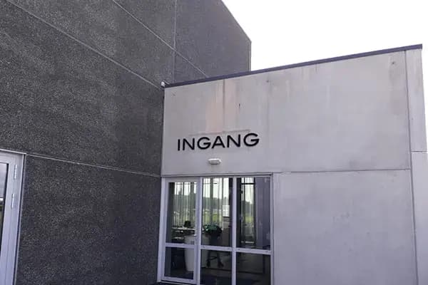 Betonnen entreegevel met zwarte losse letters "INGANG" boven een glazen toegangsdeur aan de zijkant van een bedrijfspand.