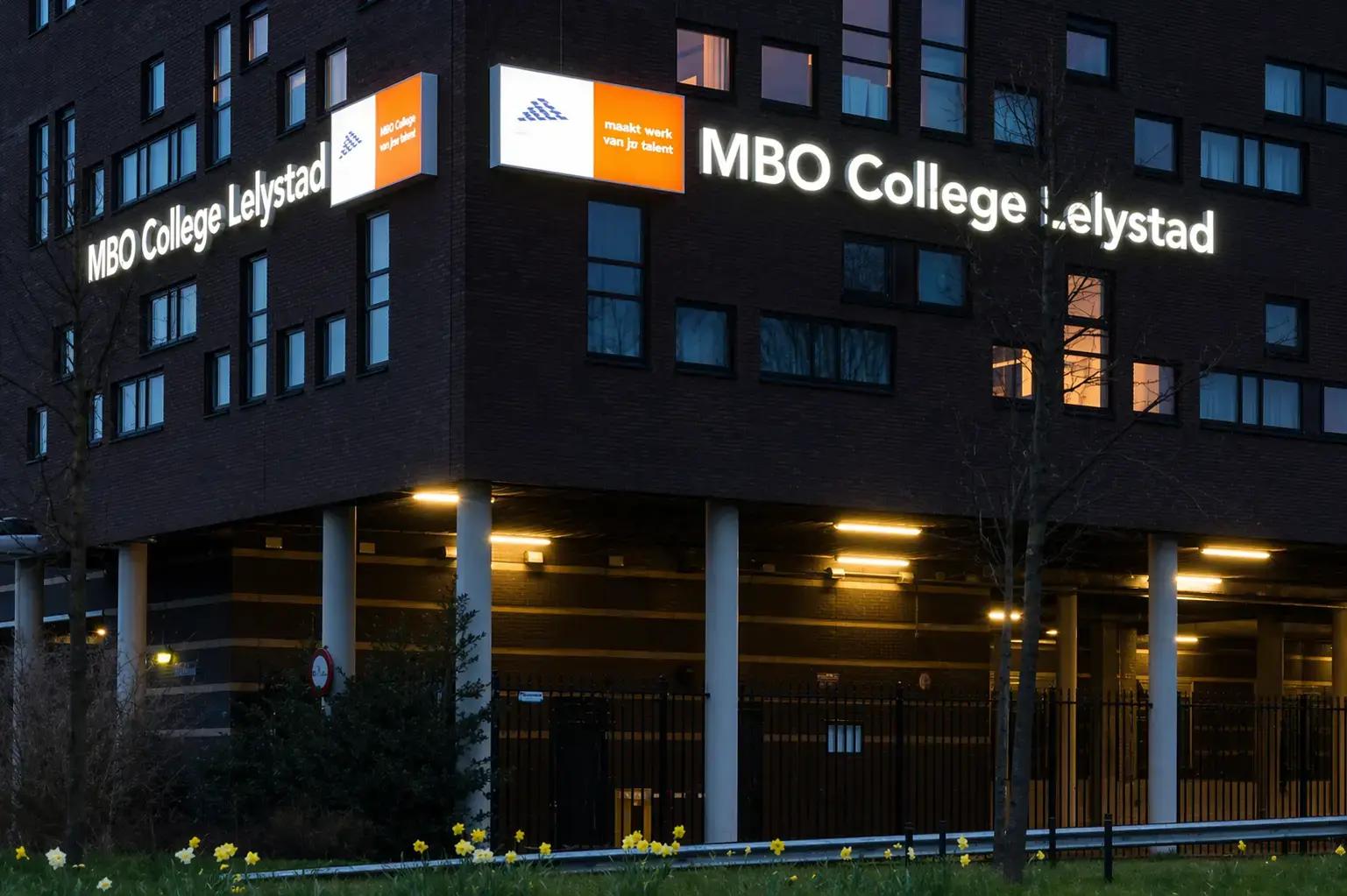 Verlichte gevelreclame met doosletters en lichtbakken voor MBO College Lelystad op een donker bakstenen schoolgebouw in de avond.