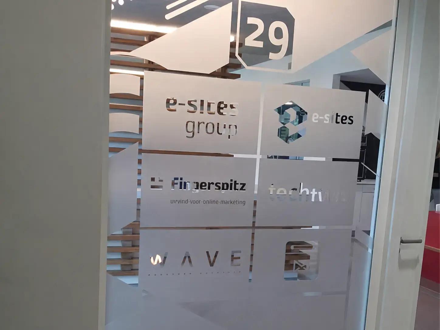 Glaswand met matte privacyfolie, uitgesneden logo’s en pijlvormen voor e-sites group, Fingerspitz en WAVE, toegepast als kantoorbelettering en wayfinding bij ruimte 29.