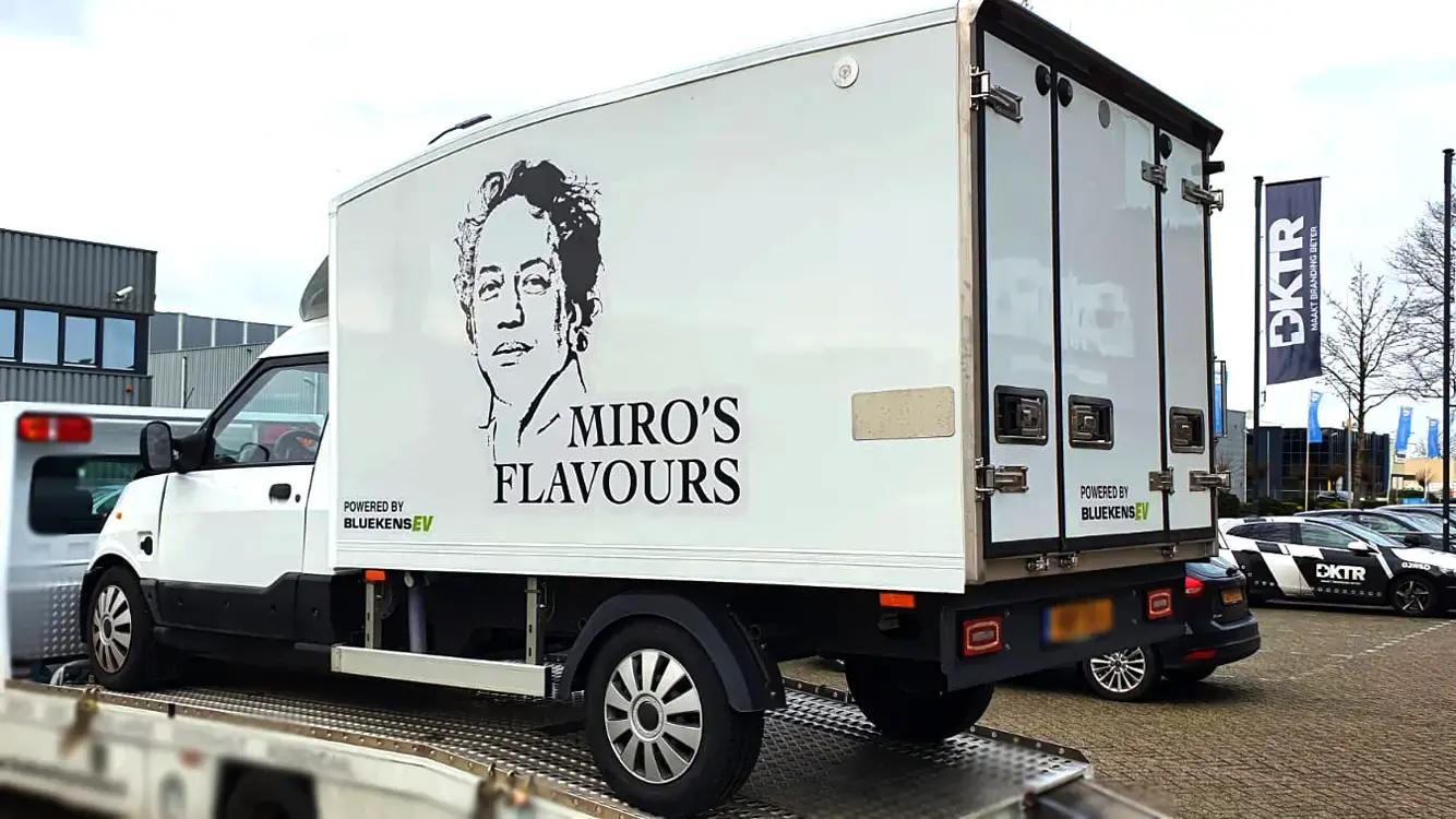 Witte bakwagen met zwarte voertuigbelettering voor Miro’s Flavours, voorzien van portretillustratie, merknaam op de laadbak en kleine sponsorvermelding op een bedrijventerrein.