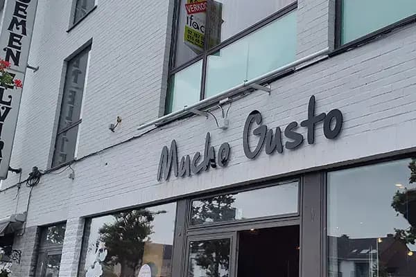 Witte bakstenen gevel met zwarte sierlijke gevelletters "Mucho Gusto" boven de glazen entree van een straatpand.